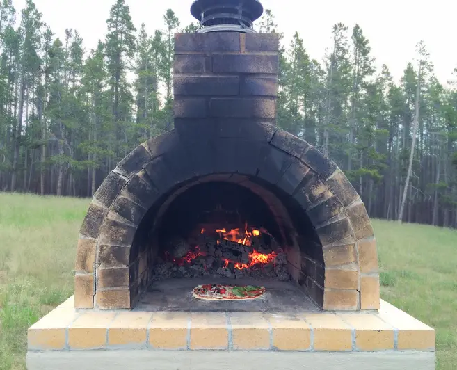 An outdoor brick oven in a grass clearing of a tree lined forest with tall pine trees. The brick oven has firewood burning and is ready to cook a pizza or loaf of bread.